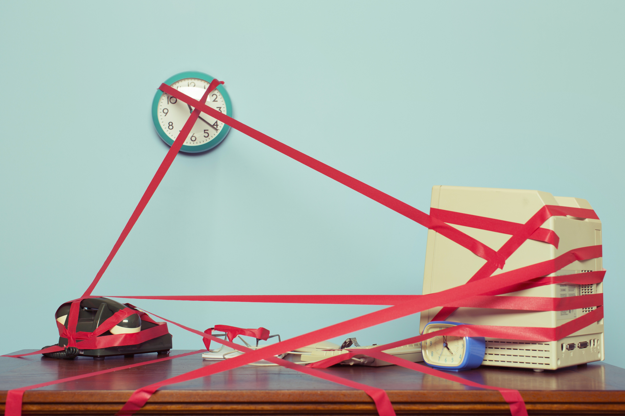 Image of a computer, phone and clock on a desk tied in red tape.