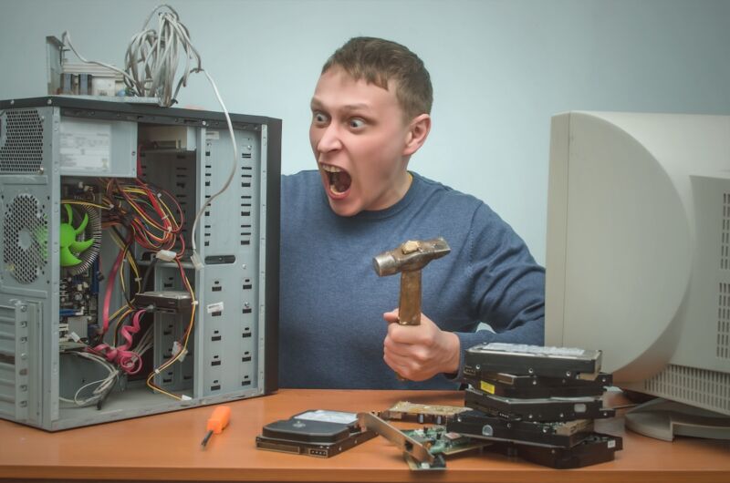 Enraged computer technician man screaming and breaking a PC with a hammer.